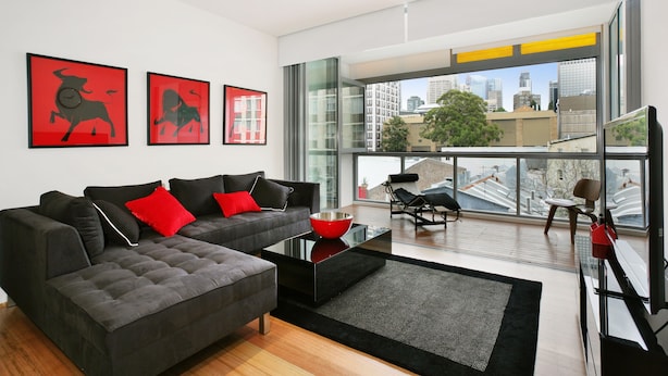 A modern living room with a grey sectional sofa, red cushions, a glass coffee table, and large windows offering a city view. - New York on Riley (Darlinghurst)