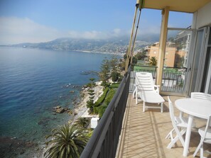 Un balcon avec des chaises blanches et une table, offrant une vue sur la côte, avec une eau d’un bleu limpide et une silhouette de ville qui se dessine à l’horizon.