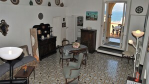 A room with a dining table, chairs, a sofa, a lamp, and a view of the sea through an open door. - Sea View Appartment for 6 Person (Gaeta)