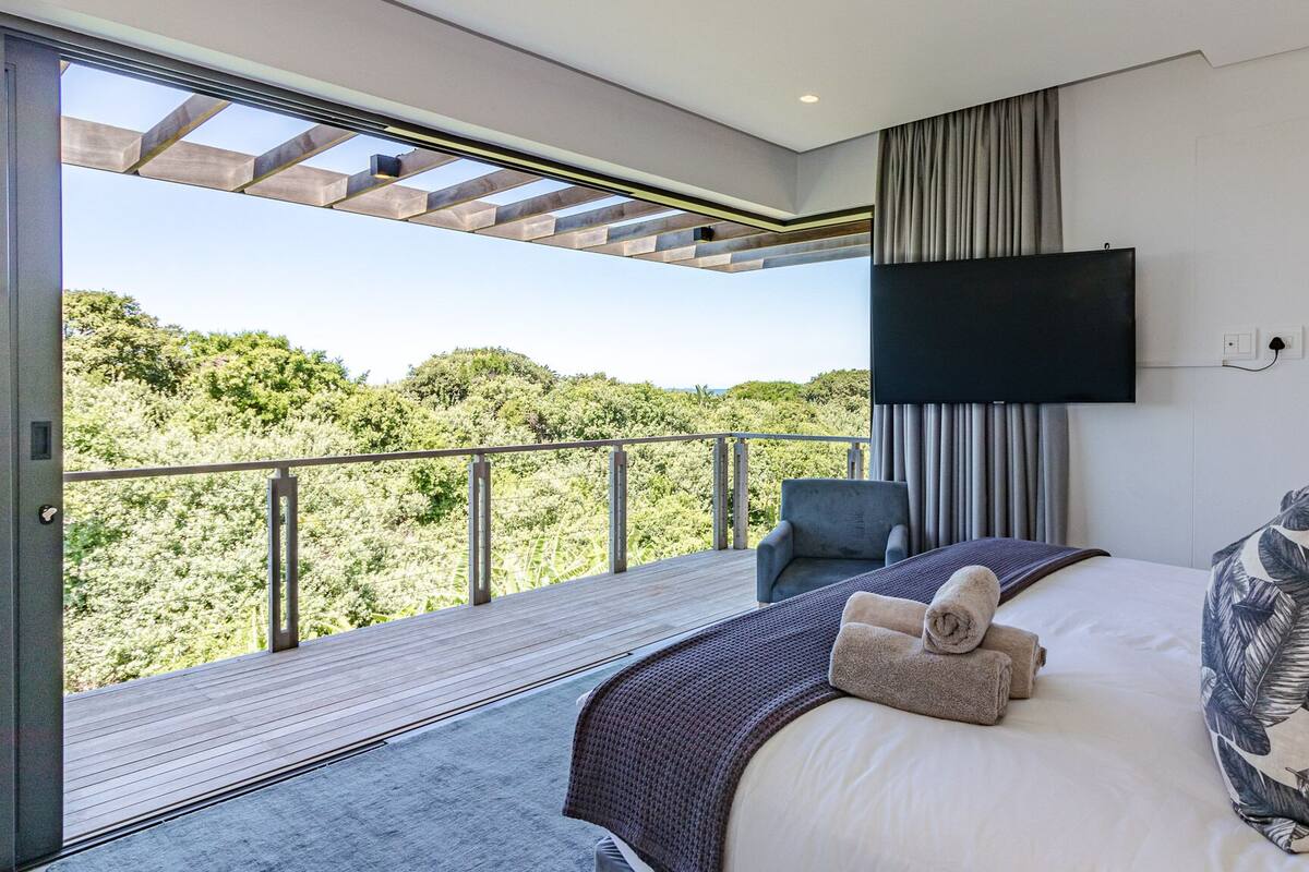 A bedroom with a large bed, a TV mounted on the wall, and a balcony with a view of greenery.