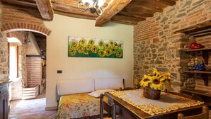 Una habitación rústica con pared de piedra, techo de madera, cama con colcha floral, mesa de comedor con un centro de girasoles y un cuadro de girasoles.