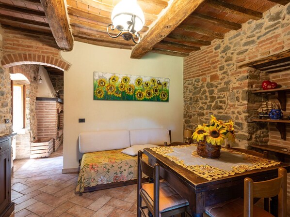 A rustic room with a stone wall, a wooden ceiling, a bed with a floral bedspread, a dining table with a sunflower centerpiece, and a painting of sunflowers.