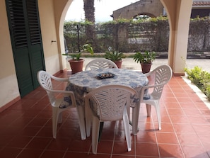 Eine Terrasse mit einem runden Tisch und weißen Stühlen, umgeben von Topfpflanzen und Blick auf ein Gebäude im Hintergrund.