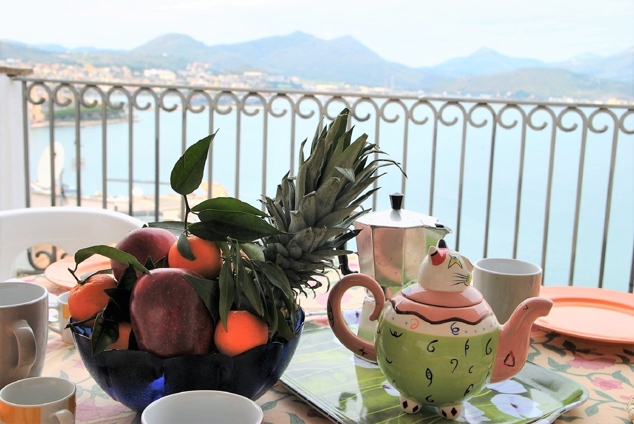 A table with a fruit bowl, a teapot, and cups on a balcony overlooking a body of water and mountains.
