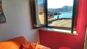 A room with a red wall, a window with a view of a coastal city, an orange sofa, and a small red stool. - Belvedere Home With sea View on the Gulf of Gaeta (Gaeta)