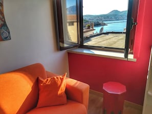 Ett rum med en röd vägg, ett fönster med utsikt över en kuststad, en orange soffa och en liten röd pall.