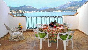 A balcony with a table set for four, white chairs, a wicker chair, and a view of a coastal town and mountains. - Belvedere Home With sea View on the Gulf of Gaeta (Gaeta)
