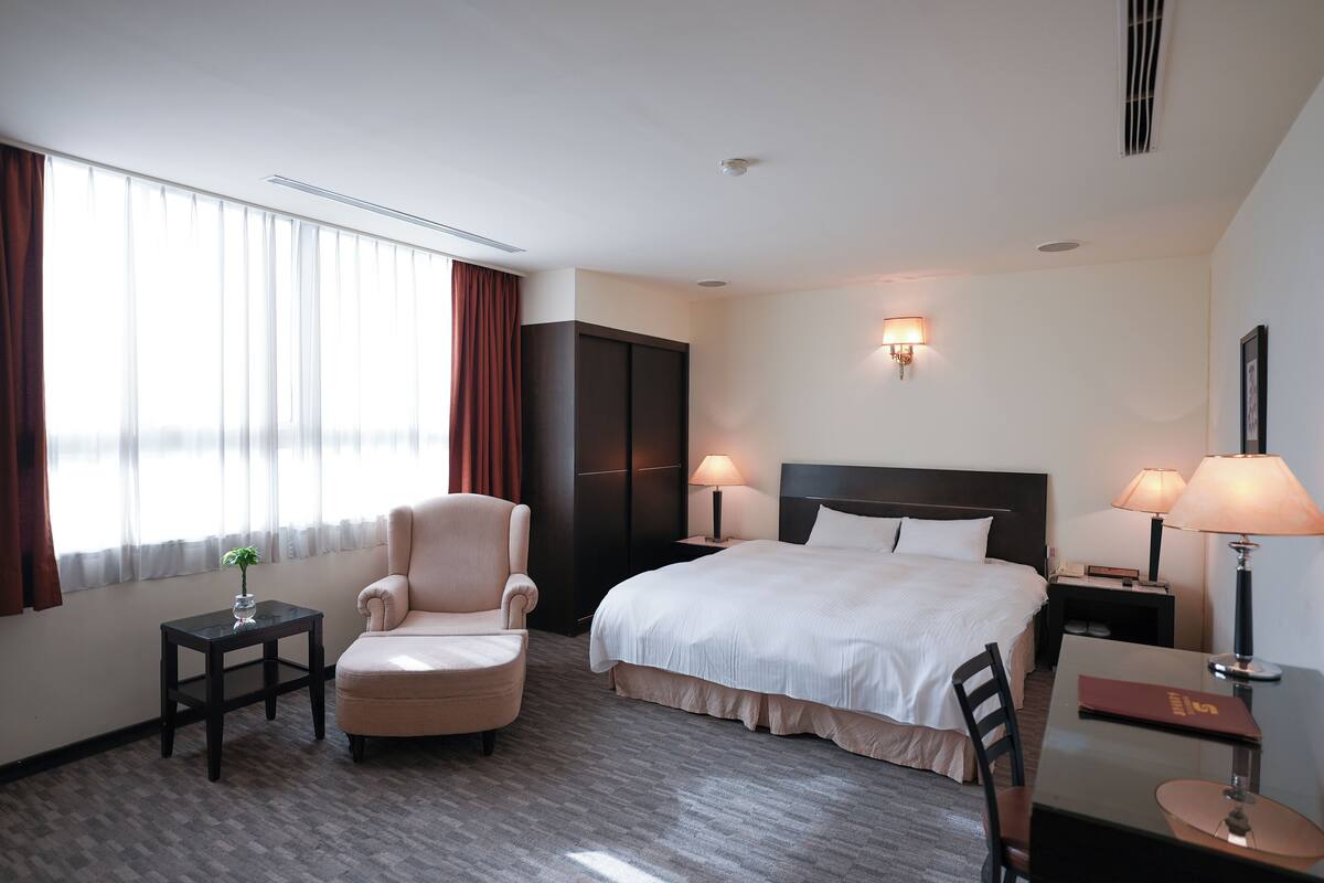 Una camera d'albergo con un letto grande, una sedia, una scrivania e un comodino con lampade.