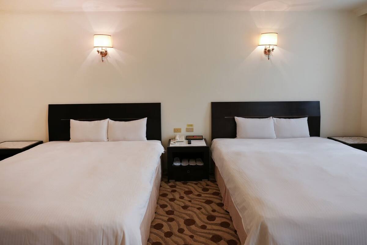 Camera d'albergo con due letti, ognuno con una testiera nera, lenzuola bianche e un piccolo comodino con una lampada e un telefono.