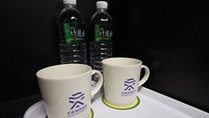 Two white mugs with blue logos on a tray, placed on a table with two bottles of water in the background. - Herkang Hotel (Taichung)