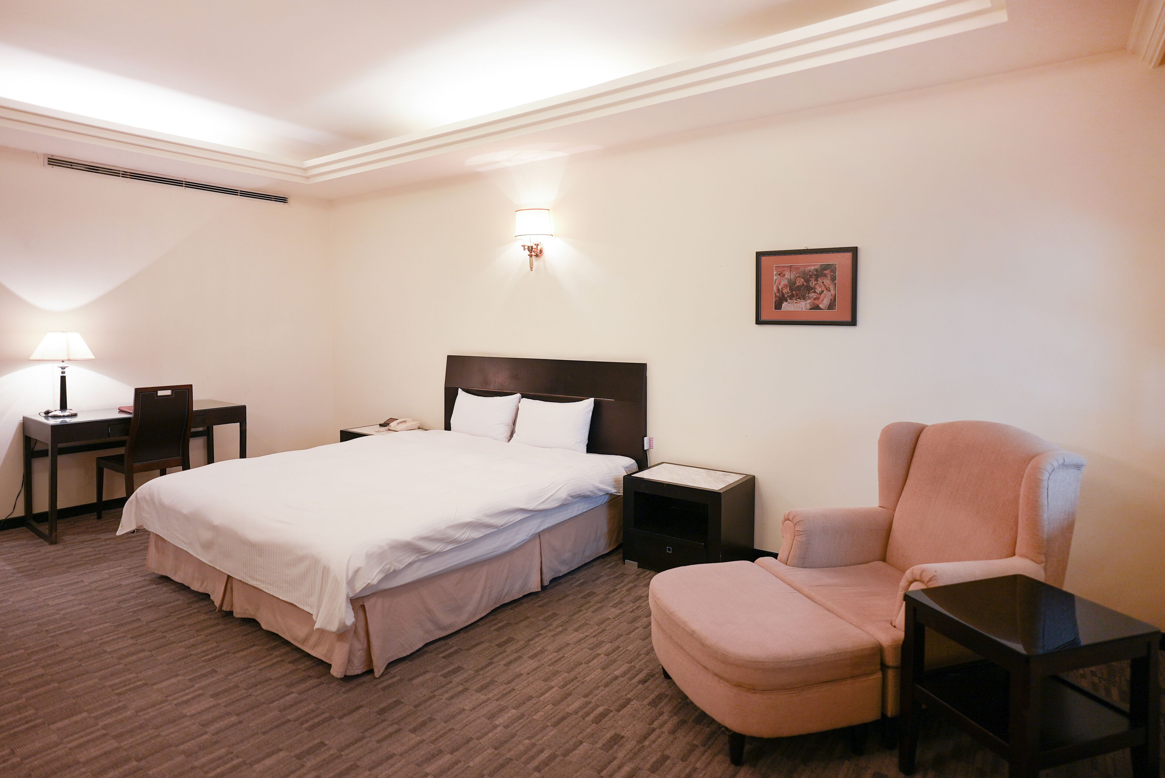 A hotel room with a large bed, a desk with a lamp, a chair, a nightstand, and a wall-mounted picture.