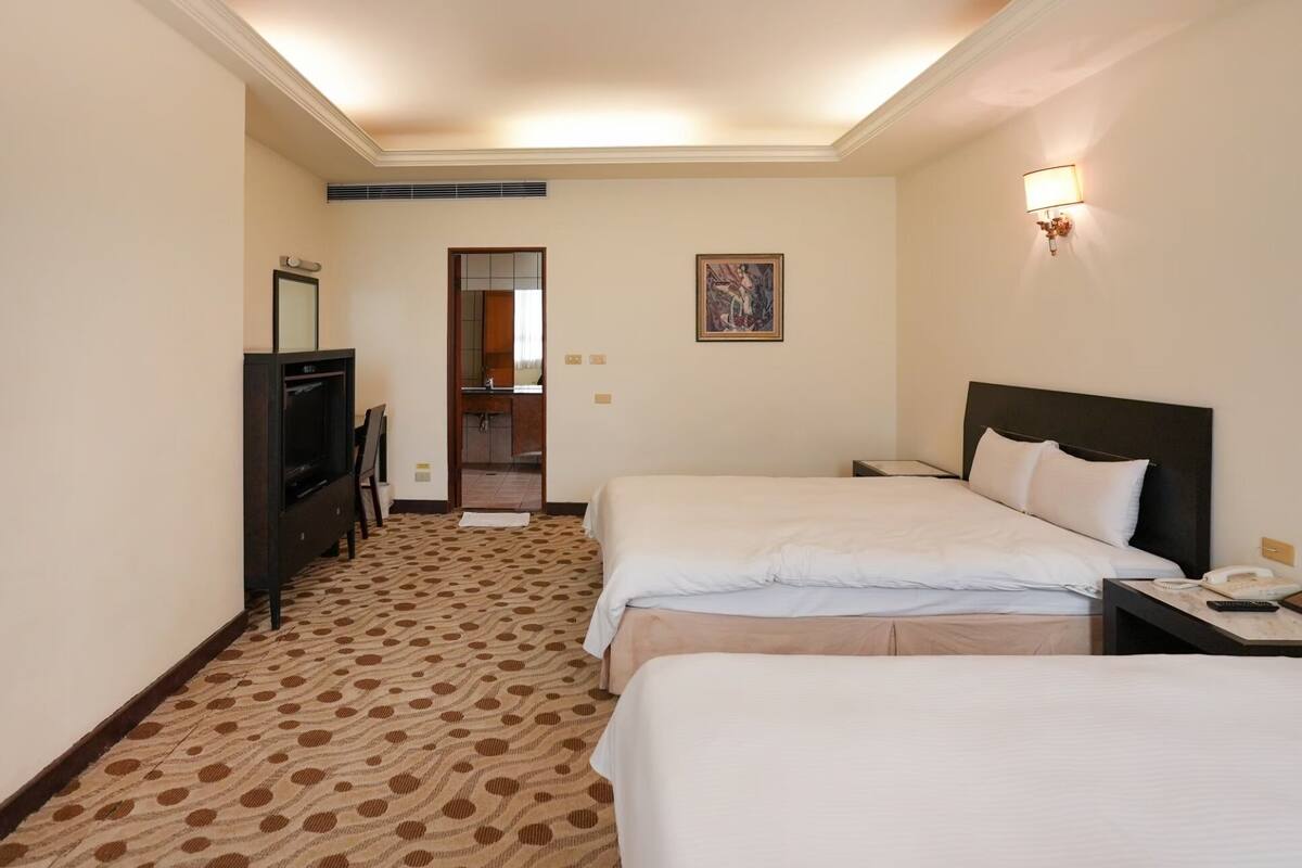 Camera d'albergo con due letti, televisione, scrivania e bagno parzialmente visibile attraverso una porta aperta.