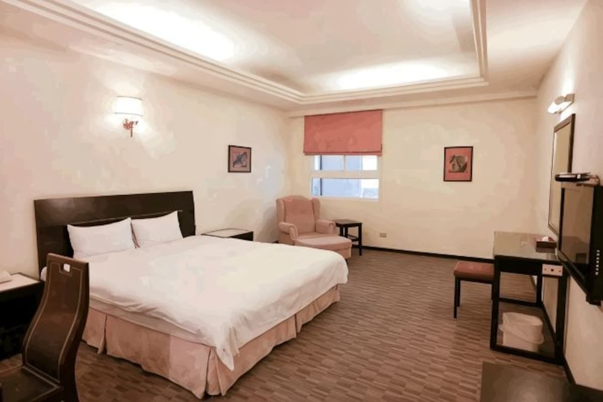 A hotel room with a large bed, a desk, a chair, a small table, and a window with a blind.