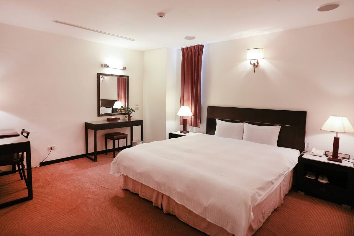 Una camera d'albergo con un letto grande, una scrivania, una sedia, un comodino con una lampada e uno specchio.