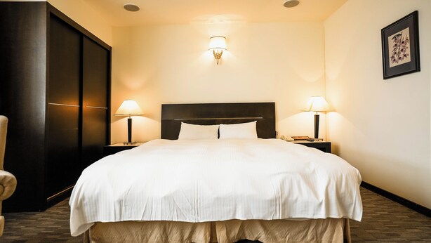 A hotel room with a large bed, two bedside lamps, a dark headboard, a wall-mounted light fixture, and a framed picture on the wall. - Herkang Hotel (Taichung)