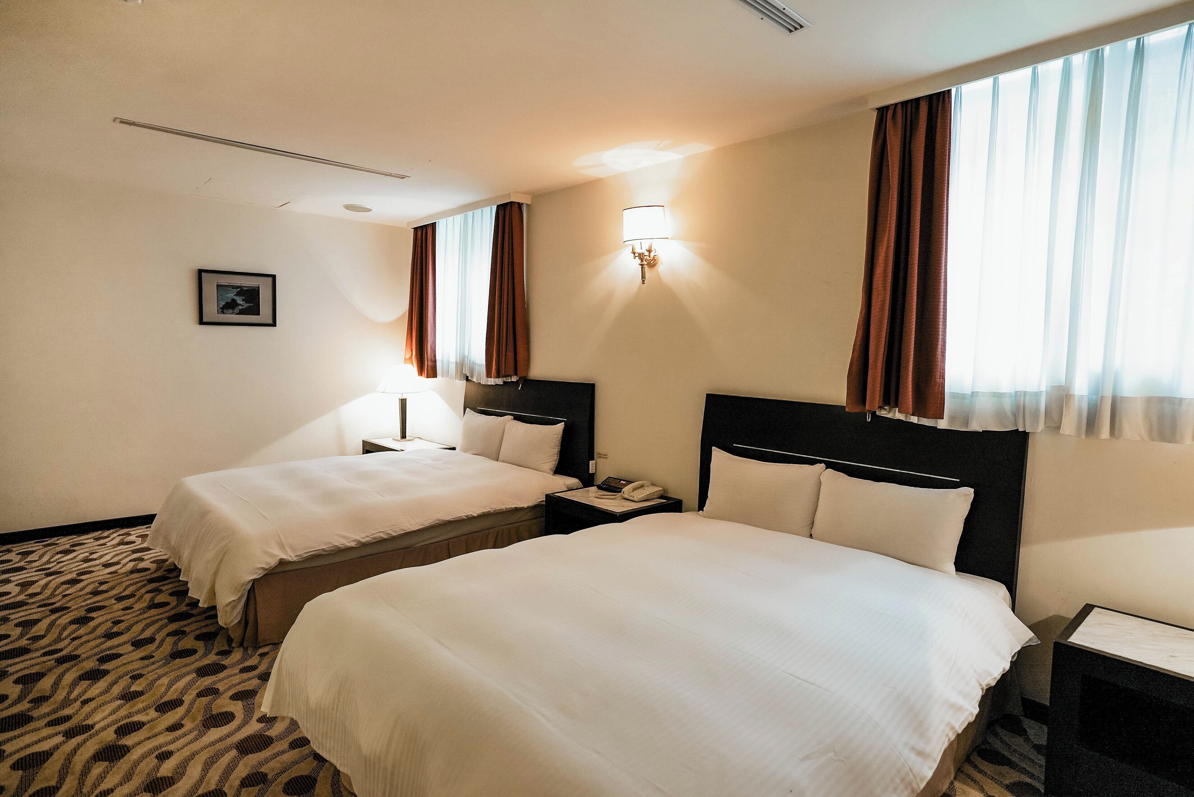 Habitación de hotel con dos camas, alfombra estampada, un cuadro enmarcado en la pared y una mesita de noche con un teléfono.