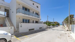 Ein modernes, zweistöckiges Gebäude mit einem Balkon im zweiten Stock, einer Treppe zum Eingang und freiem Blick auf die Straße und das Meer im Hintergrund.