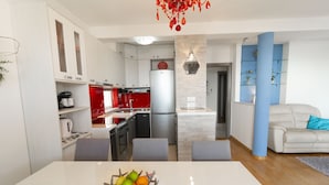 A modern kitchen with white cabinets, a red refrigerator, and a dining area with a white table and a fruit bowl. - La Storia Apartments (Makarska)