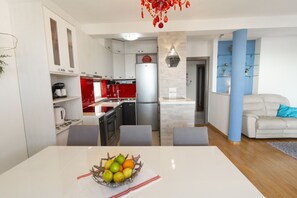 A modern kitchen with white cabinets, a red refrigerator, and a dining area with a white table and a fruit bowl. - La Storia Apartments (Makarska)