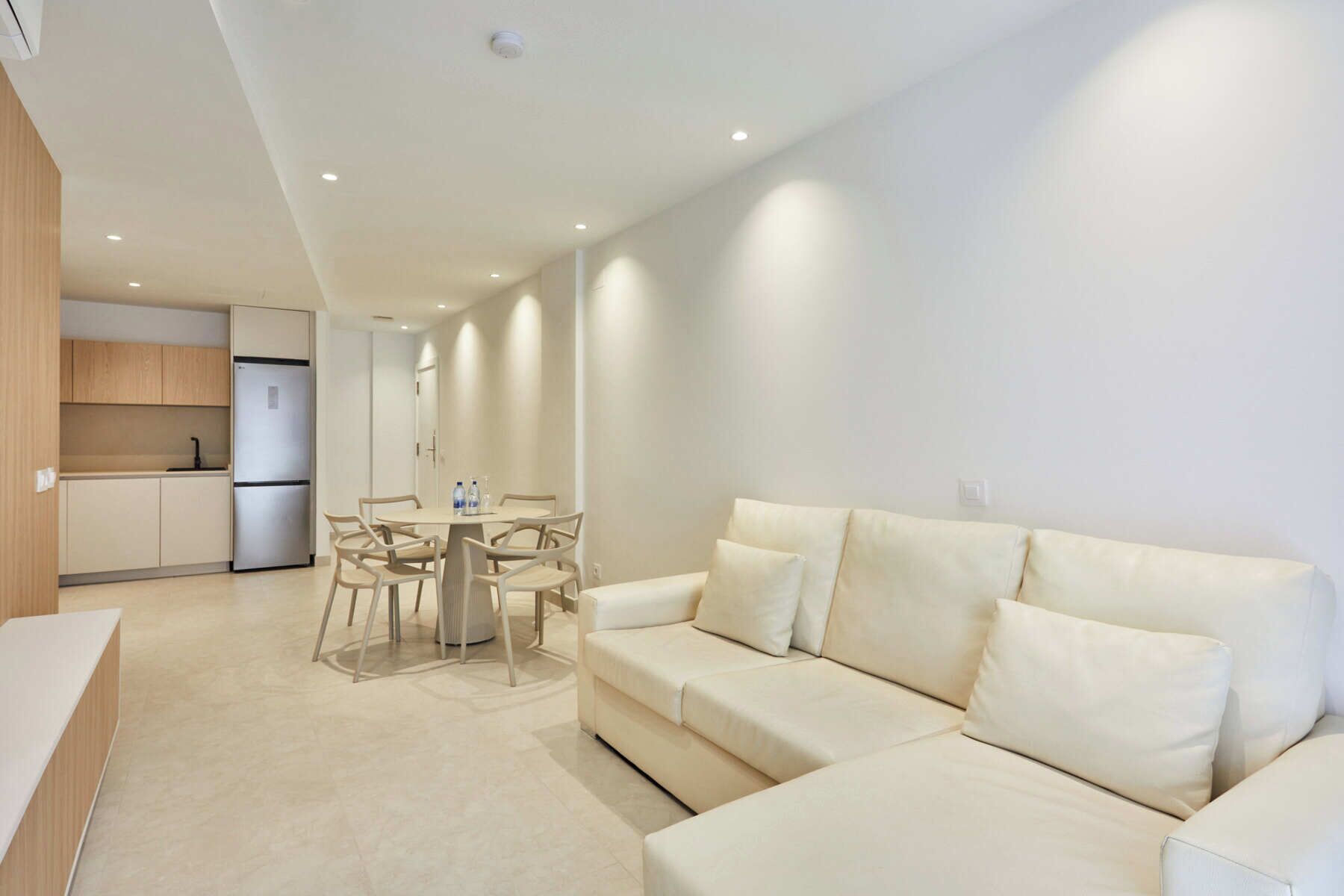 Una sala de estar moderna con un sofá color beige, una mesa de comedor con sillas, y una zona de cocina con armarios y un refrigerador.