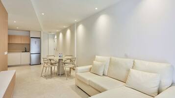 Una sala de estar moderna con un sofá color beige, una mesa de comedor con sillas, y una zona de cocina con armarios y un refrigerador.
