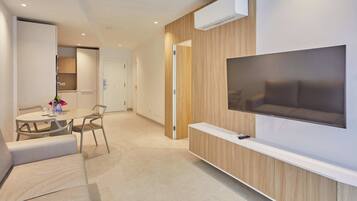 Una habitación de hotel moderna con un televisor de pantalla plana montado en un panel de madera, un área de comedor con mesa y sillas, y una puerta que conduce a un baño.