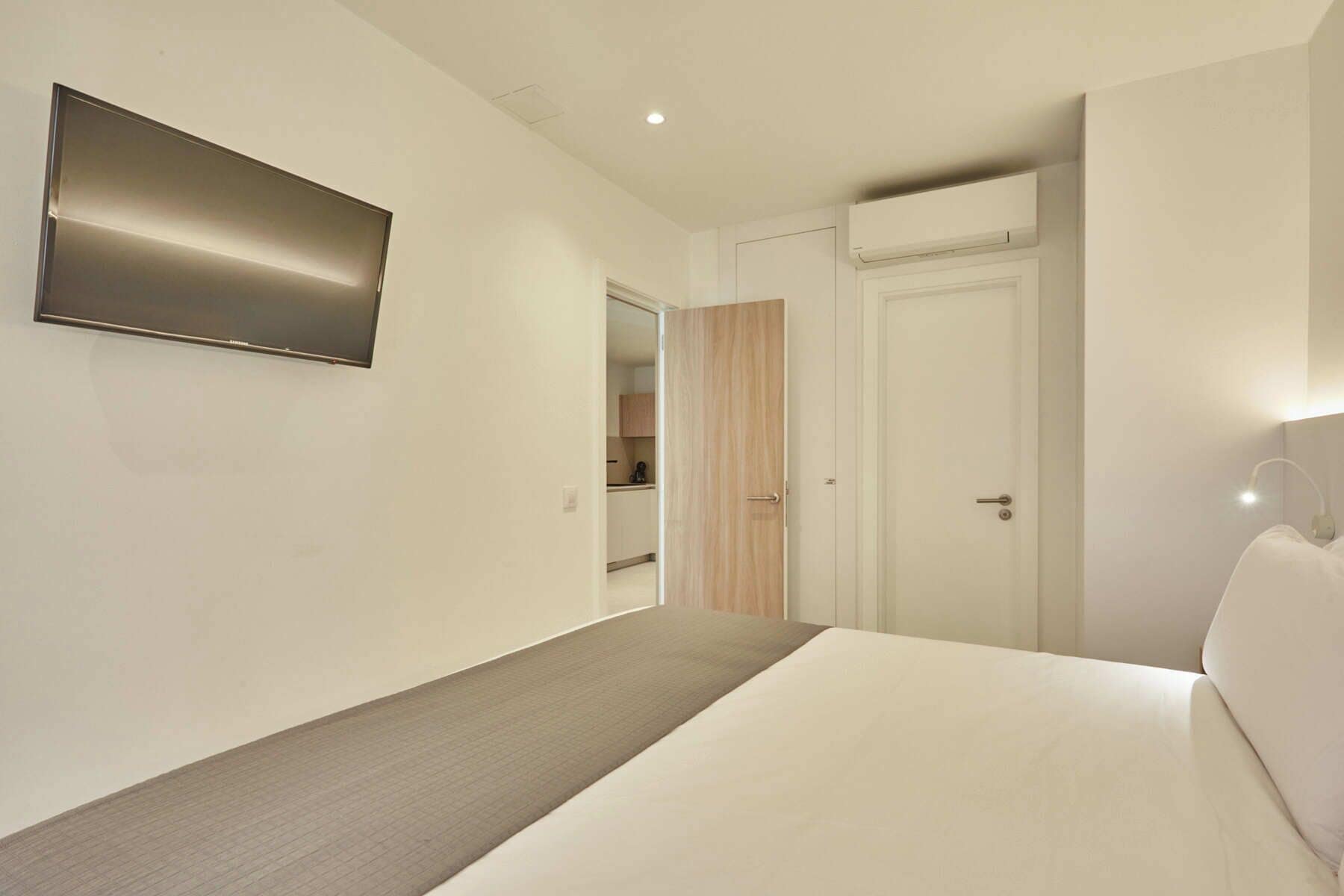 A hotel room with a bed, a flat-screen TV mounted on the wall, a door leading to a bathroom, and a bedside table with a lamp.