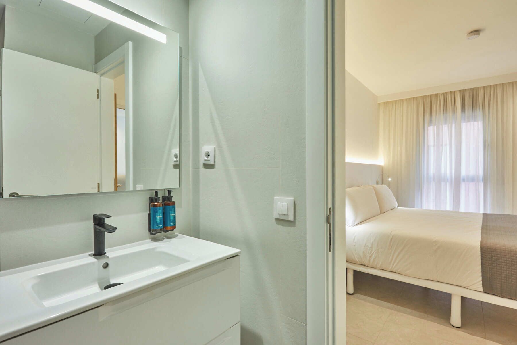 Habitación de hotel con lavamanos blanco, espejo y artículos de aseo. Se observa una cama con ropa blanca y una ventana con cortinas.