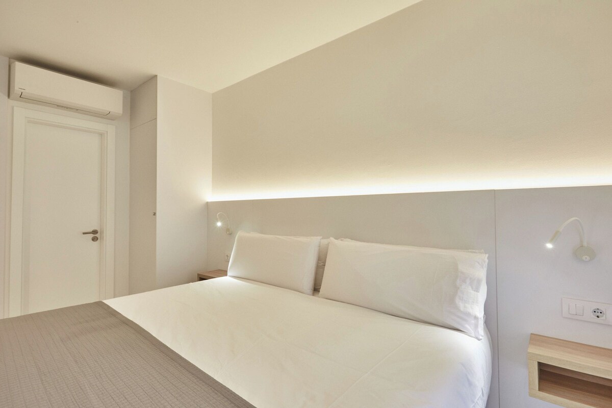 A bedroom with a bed, a door, a nightstand, and a wall-mounted light fixture.