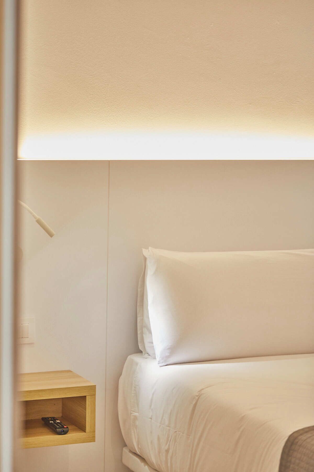 A bed with white bedding, a bedside table with a remote control, and a wall-mounted light fixture.