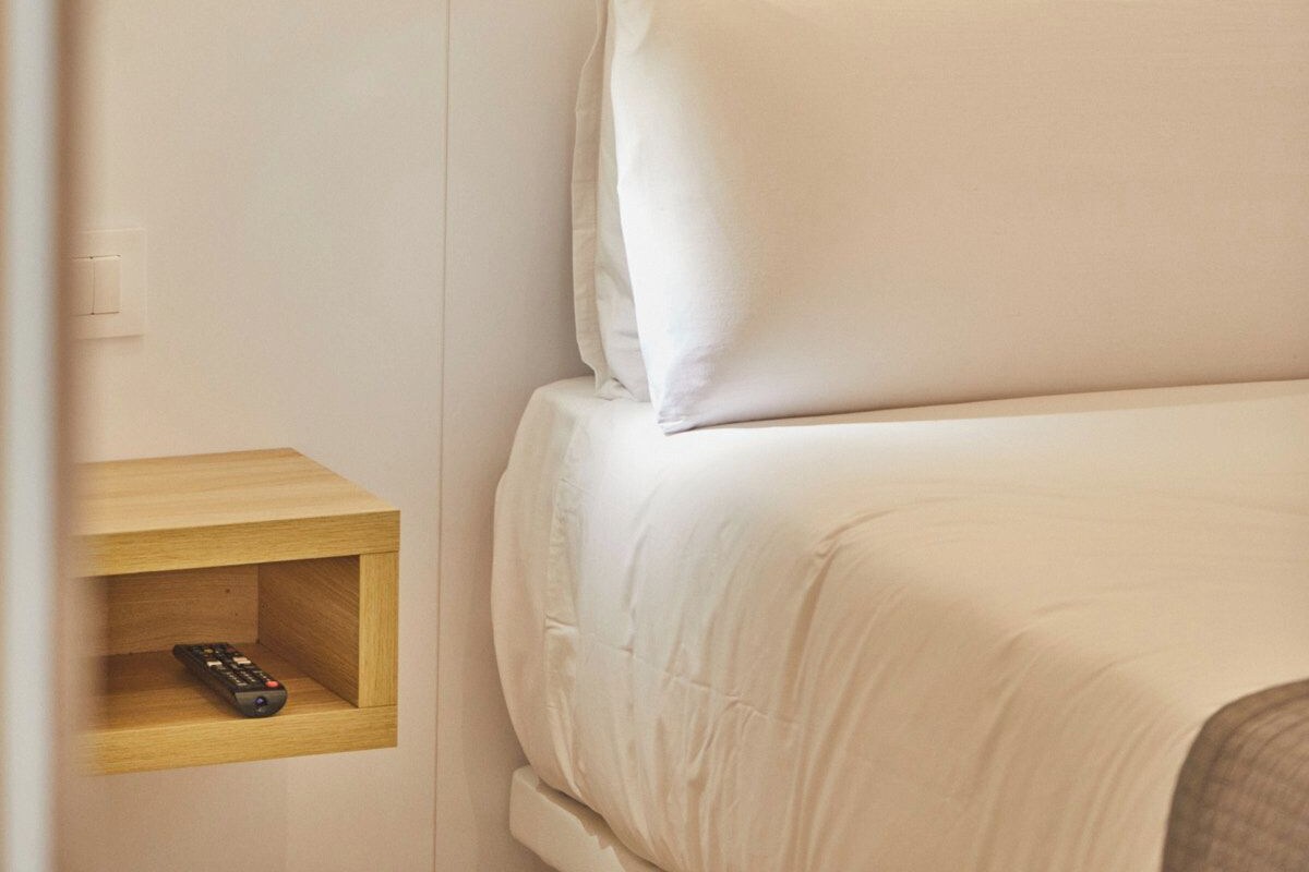 Un lit avec une literie blanche, une table de chevet avec une télécommande et un luminaire fixé au mur.