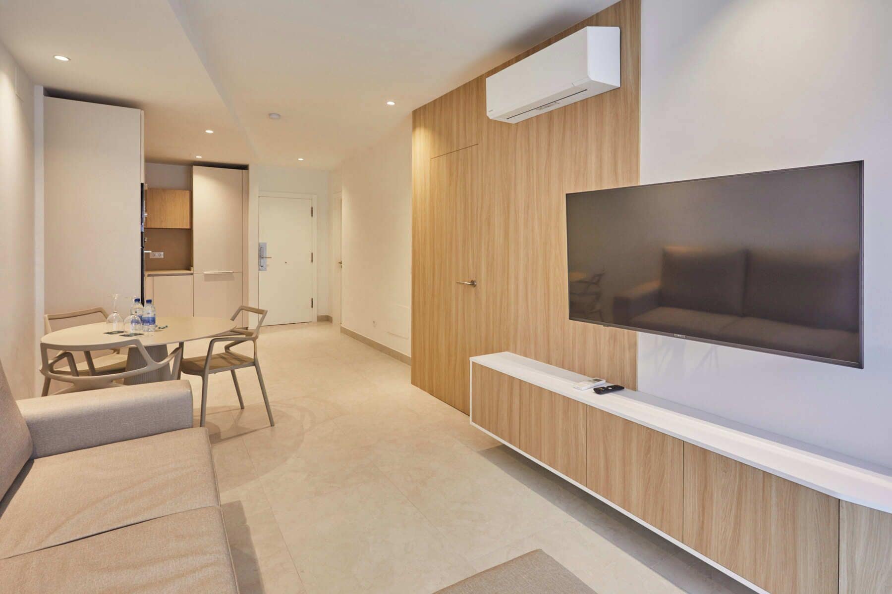 Una habitación de hotel moderna con un televisor de pantalla plana montado en un panel de madera, una mesita con sillas y un sofá.