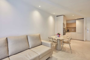 A modern living room with a beige sofa, a round dining table, and a kitchen in the background. - Silken Atlantis Playa – Holiday Homes (Las Palmas de Gran Canaria)