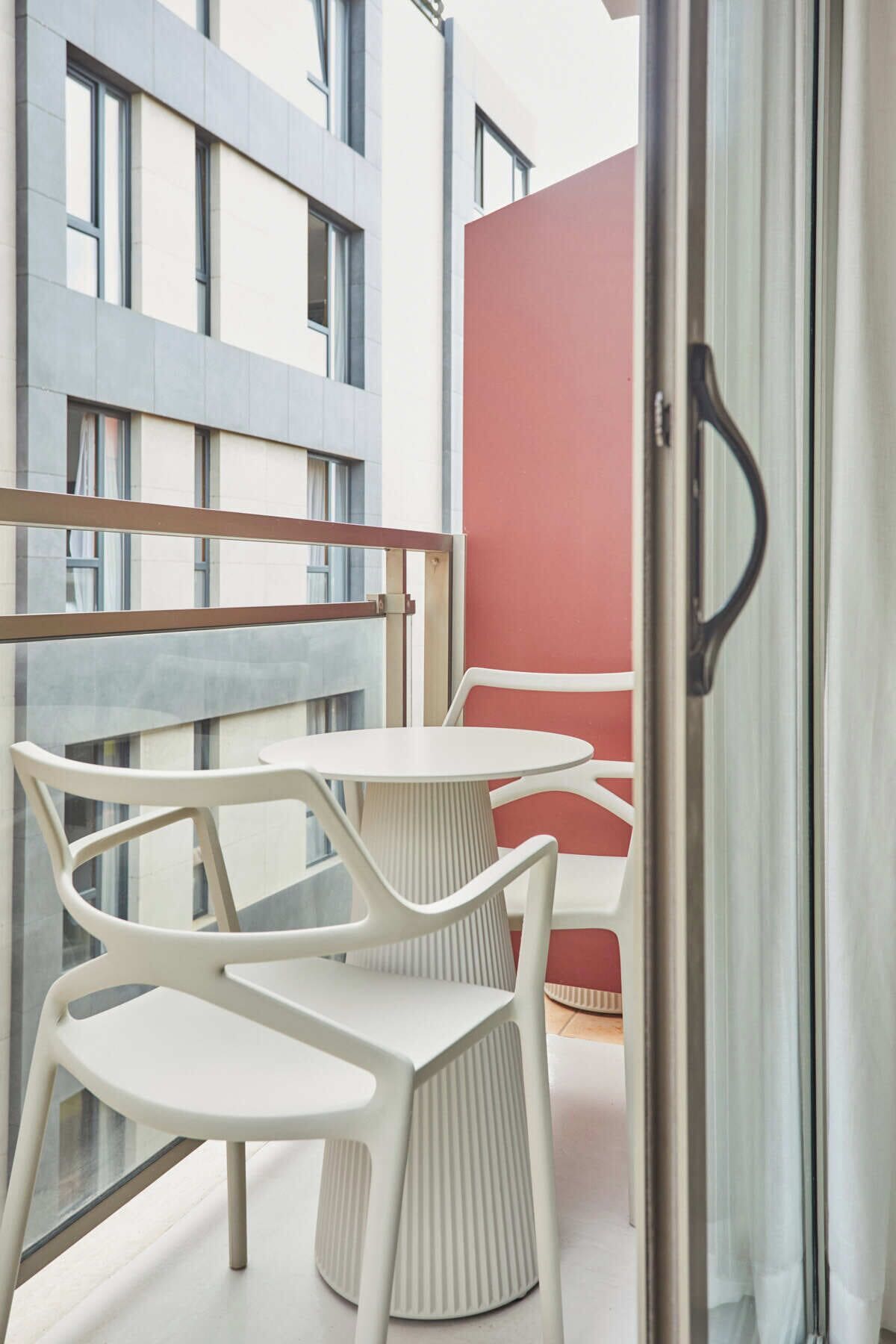 Ein Balkon mit zwei weißen Plastikstühlen und einem Tisch, einer roten Wand und einer Glastür.