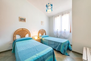 A bedroom with two single beds, a bedside table with a lamp, a window with curtains, and a wall-mounted picture.