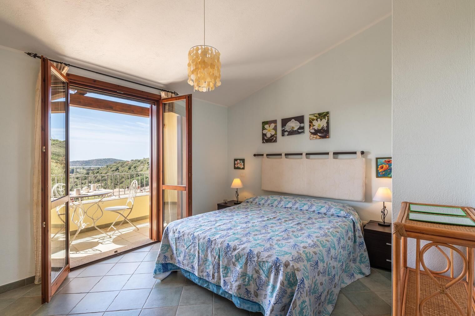Un dormitorio con cama, mesita de noche, lámpara y vistas a un balcón con mesa y sillas.