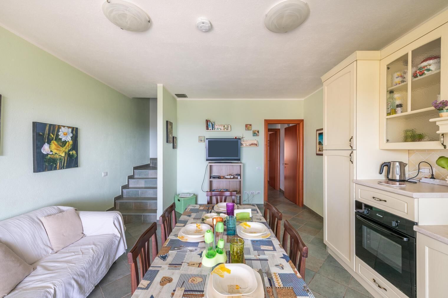 A dining area with a table set for four, a sofa, a kitchen with an oven, and a staircase leading to another room.