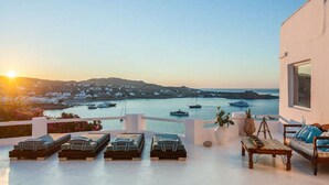 A terrace with sun loungers, a wooden table, and a view of boats and a coastline at sunset. - Villa Panoramic Mykonos Near Nammos (Mykonos)