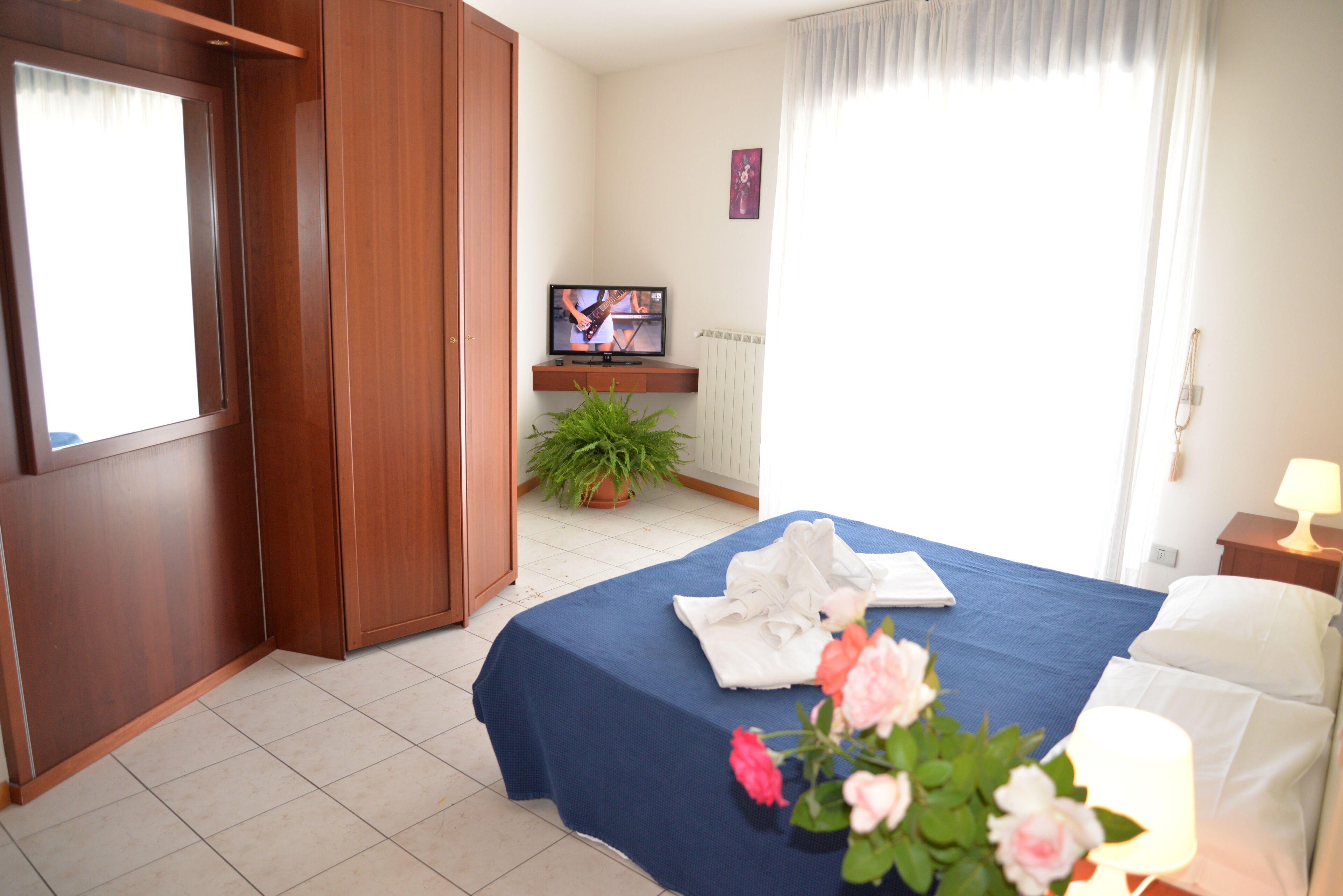Una habitación de hotel con cama, televisión, planta en maceta y ventana con cortinas.