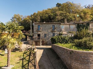 A stone house with a small balcony, surrounded by lush greenery and a palm tree.