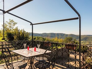A covered outdoor patio with a table and chairs, a view of the countryside, and a metal railing.