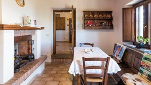 A rustic kitchen with a brick fireplace, wooden beams, a dining table with chairs, and a shelf with kitchenware.