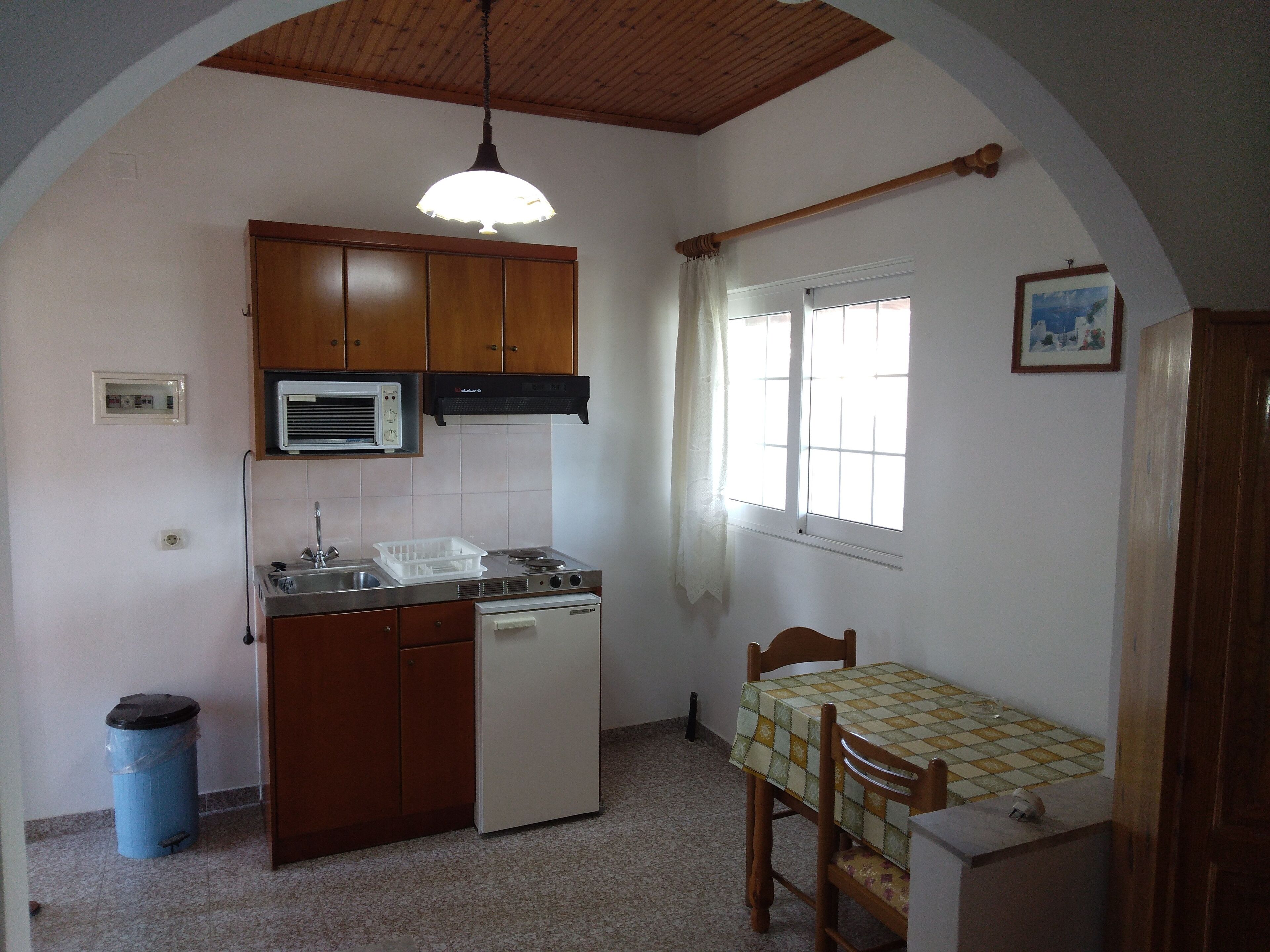 Cocina compacta con armarios de madera, microondas integrado, un lavamanos y una pequeña mesa de comedor con sillas.