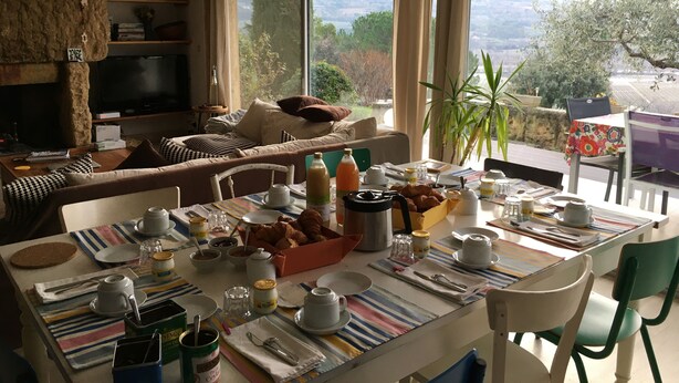 Et spiseområde med et bord dækket til morgenmad, en sofa med puder, et fjernsyn og udsigt til grøn natur.