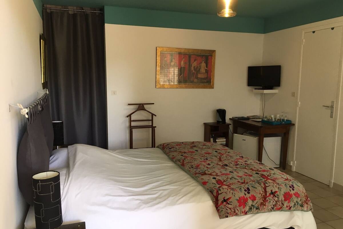 A bedroom with a bed, a desk, a television, a painting, and a lamp.