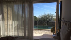 Et soveværelse med seng, et vindue med let gardin, udsigt til en pool og en balkon med en stol.