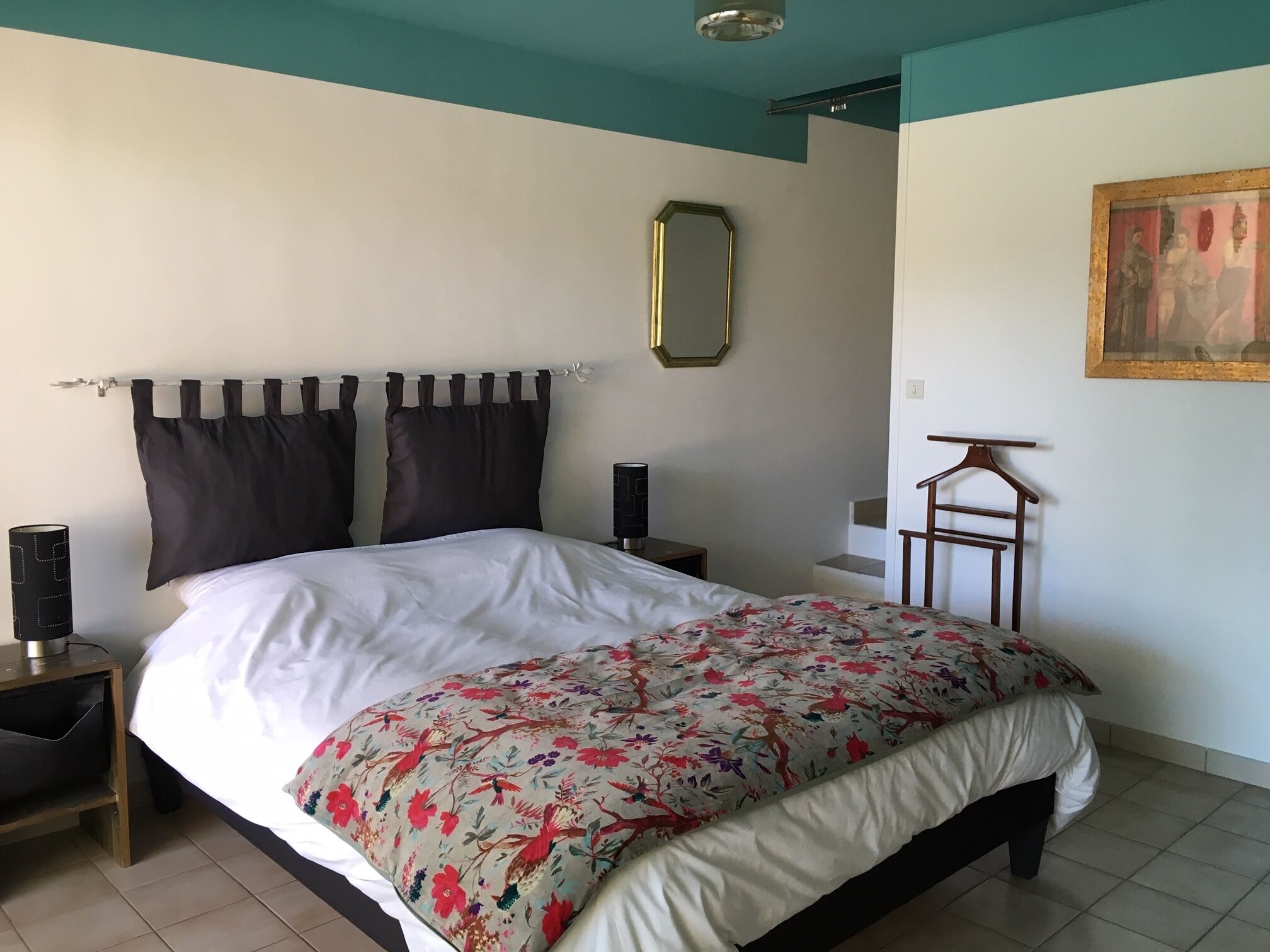 Een slaapkamer met een bed, nachtkastje, kapstok, spiegel en een schilderij aan de muur.