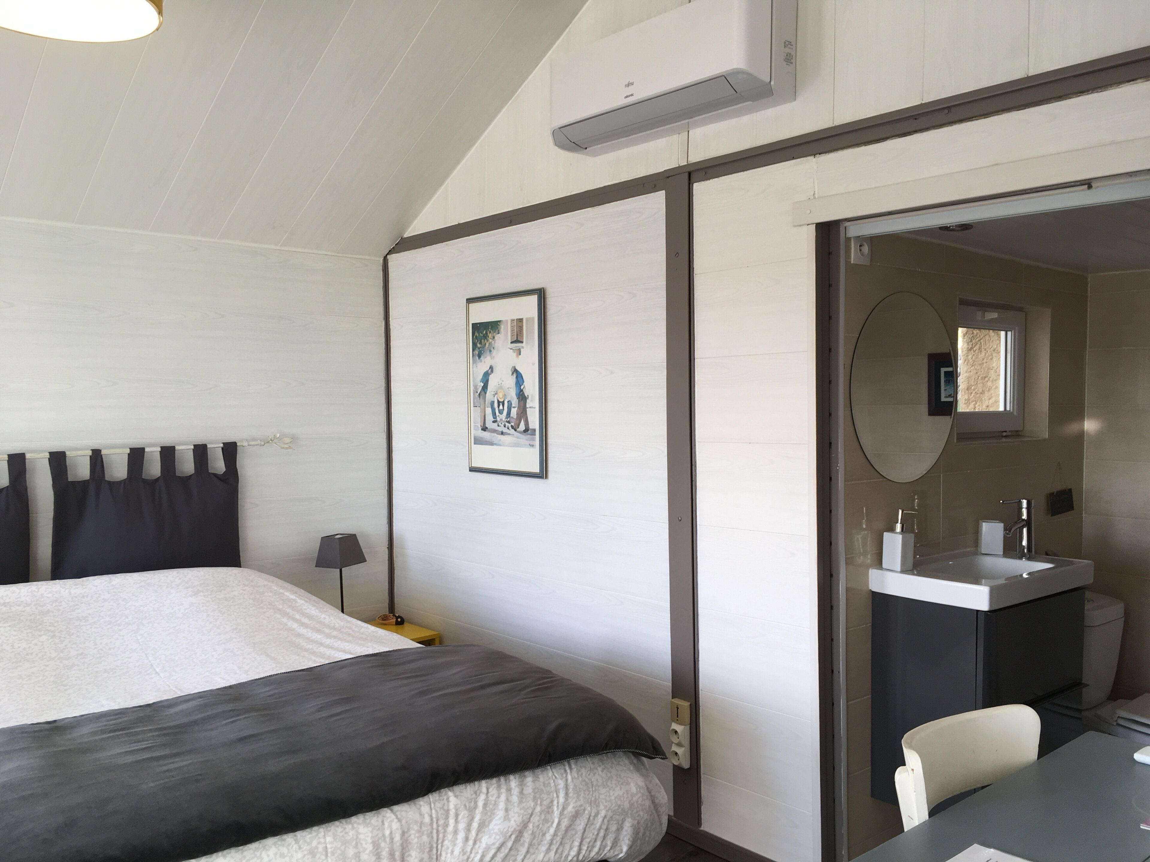 A bedroom with a bed, a bedside lamp, a framed picture on the wall, and a bathroom with a sink and mirror.