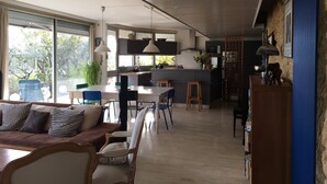Et moderne køkken med en central ø, spiseområde med sofa og stole, samt udsigt over naturen gennem store vinduer.