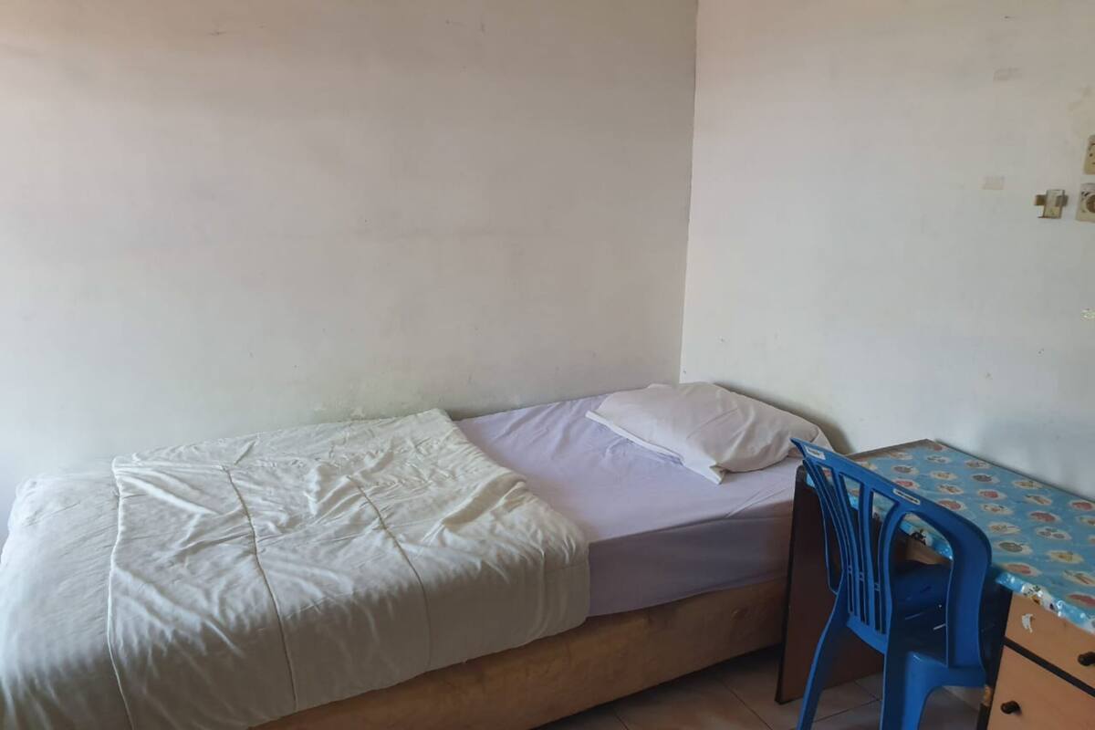 A small, single-bed room with a blue plastic chair and a table with a patterned tablecloth.